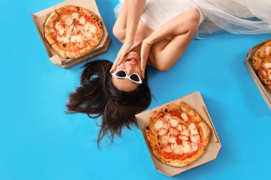 Young woman in white dress with tasty pizza lying on blue background, top view