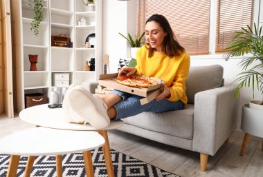 Evdeki kanepede oturan bir kutu lezzetli pizzayla güzel bir kadın.
