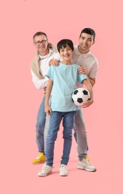 Mutlu küçük çocuk, babası ve dedesiyle birlikte pembe arka planda futbol topu tutuyor.