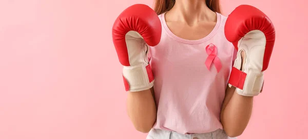 Young woman in boxing gloves and with pink ribbon on color background. Breast cancer awareness concept