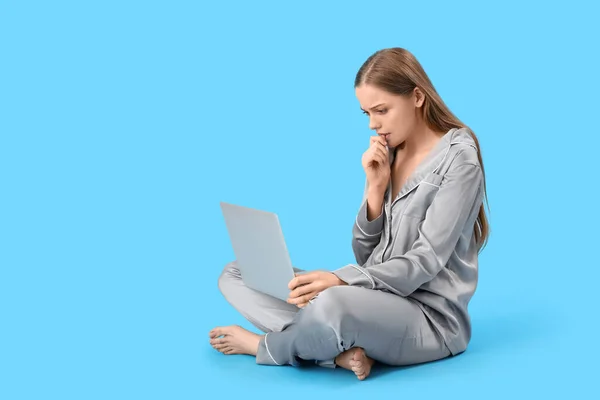 Young woman in pajamas with laptop biting nails on blue background