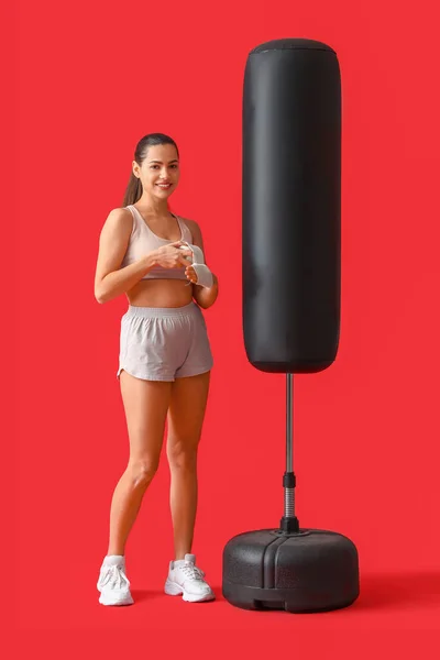 Female boxer applying wrist wraps and punching bag on red background