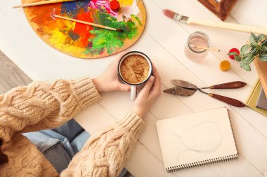 Beyaz ahşap masada bir fincan kahveli kadın sanatçı, yakın plan.