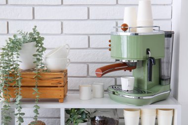 Modern coffee machine with cups and houseplants on shelving unit near white brick wall clipart