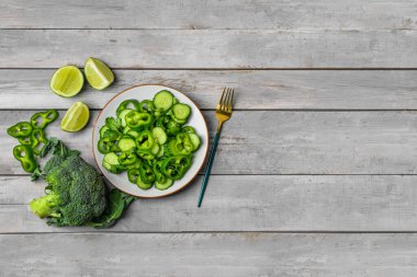 Yeşil sebzeli bir tabak salata ve gri ahşap arka planda malzemeler.