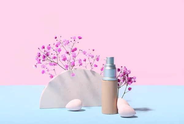 Bottle of makeup foundation, decorative plaster figure and gypsophila flowers on blue table against pink background
