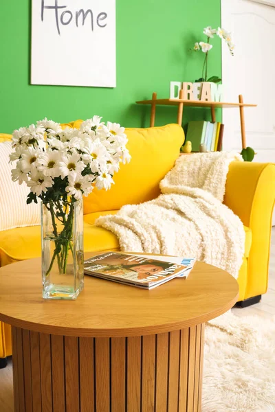 Vase with flowers and magazines on table near yellow sofa in living room