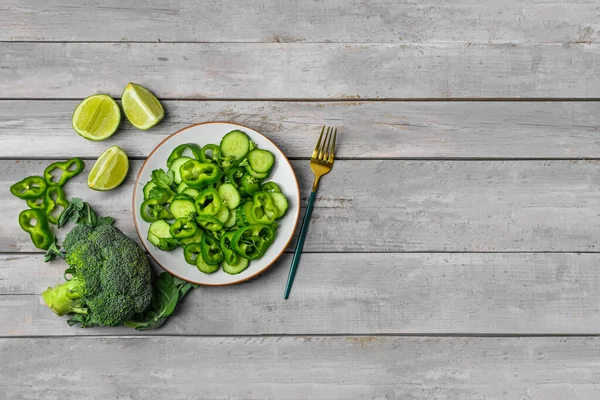Yeşil sebzeli bir tabak salata ve gri ahşap arka planda malzemeler.