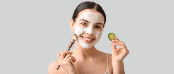 Beautiful young woman with cucumber slice applying facial mask on light background