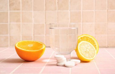 Bir bardak su, C vitamini tablet ve pembe kartonlu masada meyveler.