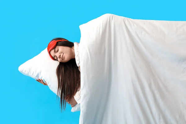 Young woman with sleeping mask, pillow and blanket on blue background