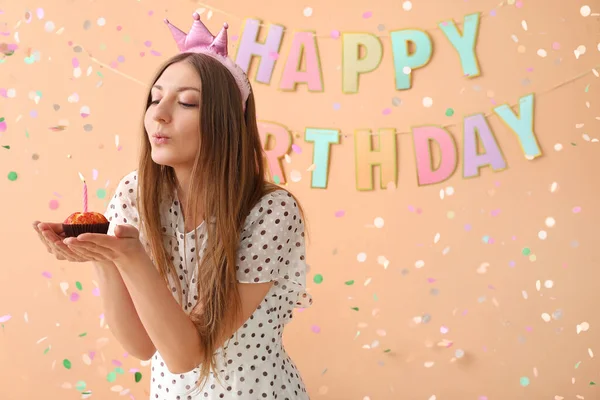 Happy young woman with Birthday muffin making wish on beige background