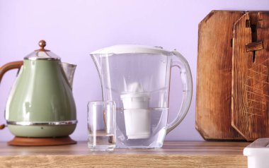 Su filtresi sürahi, cam ve su ısıtıcısı mutfak tezgahında, leylak duvarın yanında.