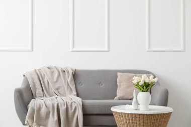 Yastıklı rahat gri kanepe ve beyaz duvarın yanındaki masada lale çiçekleri olan vazo.