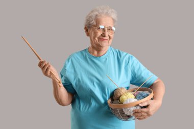 Gri arka planda örgü iplikleri ve iğneleri olan yaşlı bir kadın.