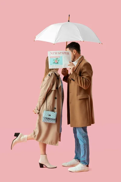 在粉色背景的报纸后面 一对年轻夫妇带着雨伞亲吻 — 图库照片