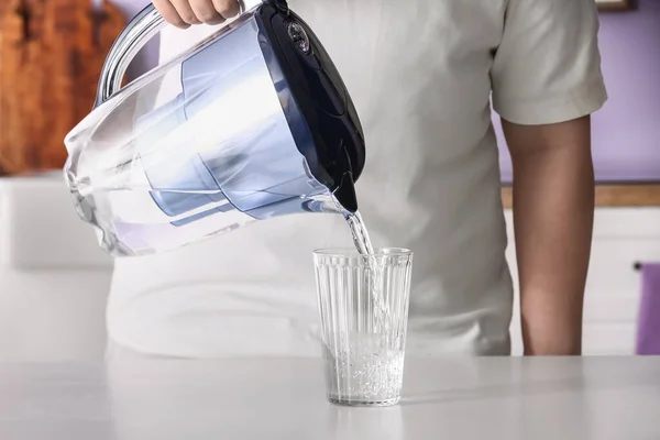 Man pouring water from filter jug into glass on table in kitchen, closeup