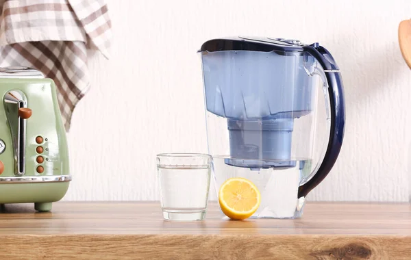 Modern filter jug, glass of water and lemon on kitchen counter