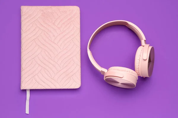 Notebook with modern pink headphones on purple background