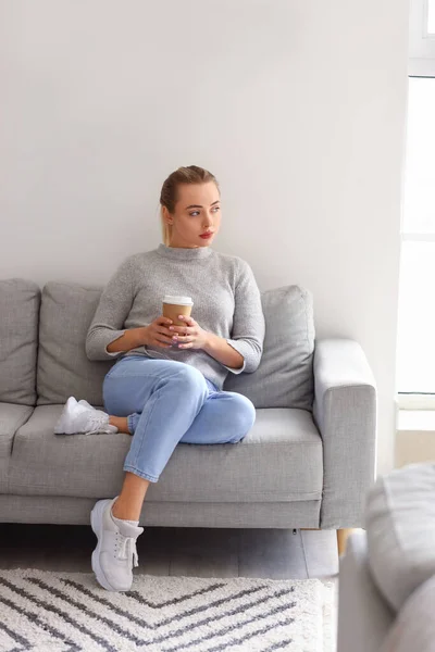 Thoughtful young woman with cup of coffee sitting on sofa at home