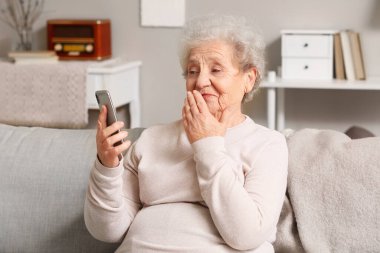 Cep telefonlu yaşlı kadın evdeki kanepede sohbet ediyor.