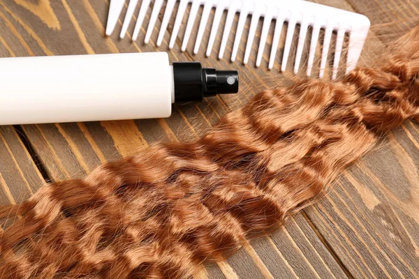 Curly ginger hair with comb and spray on wooden background, closeup