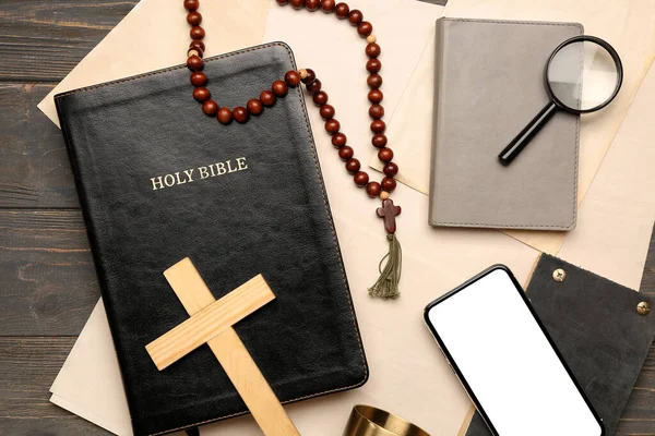 Holy Bible with prayer beads, cross, mobile phone, notebooks and paper sheets on dark wooden background