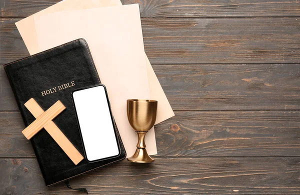 Holy Bible with cross, mobile phone, wine cup and paper sheets on dark wooden background