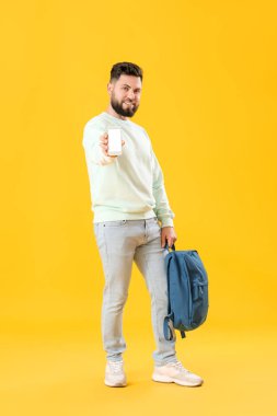 Young bearded man with backpack and mobile phone on yellow background