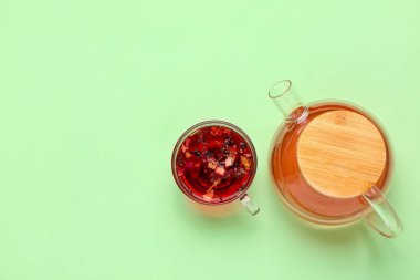 Yeşil zemin üzerinde bir fincan meyve çayı olan şeffaf çaydanlık.