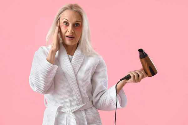 Surprised mature woman with hair dryer on pink background