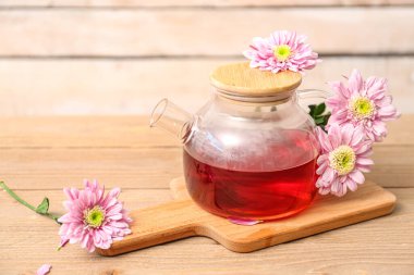 Duvarın yanındaki ahşap masada krizantem taşıyan şeffaf çaydanlık.