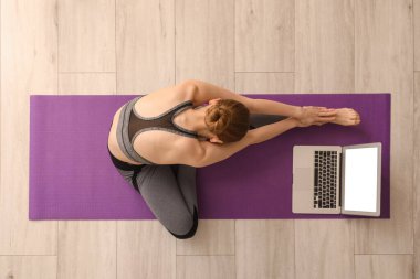 Dizüstü bilgisayarlı genç bir kadın spor salonunda yoga yapıyor.