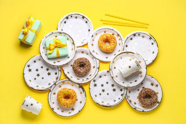Sarı zemin üzerinde doğum günü hediyeleri ve çörekleri olan tek kullanımlık kağıt masa takımı.