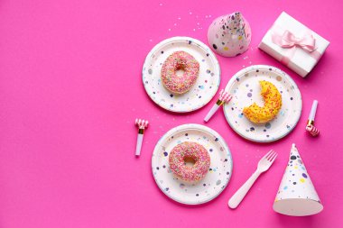 Tek kullanımlık kağıt tabaklar, donutlar, parti üfleyicileri ve pembe arka planda şapka.