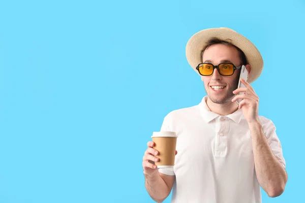 Young man with cup of coffee talking by mobile phone on blue background
