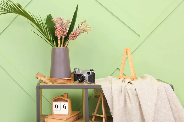 Vase with baby pineapples, palm leaves and photo camera on shelf near green wall