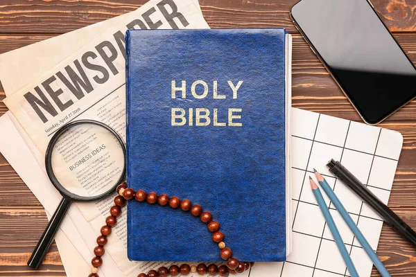 Holy Bible with prayer beads, mobile phone, magnifier, newspaper and notebook on wooden background