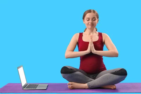 stock image Pregnant young woman with laptop doing yoga on blue background