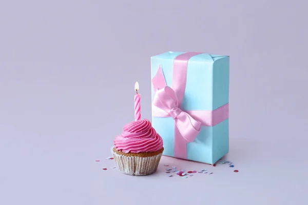 Tasty cupcake with birthday candle and gift box on lilac background