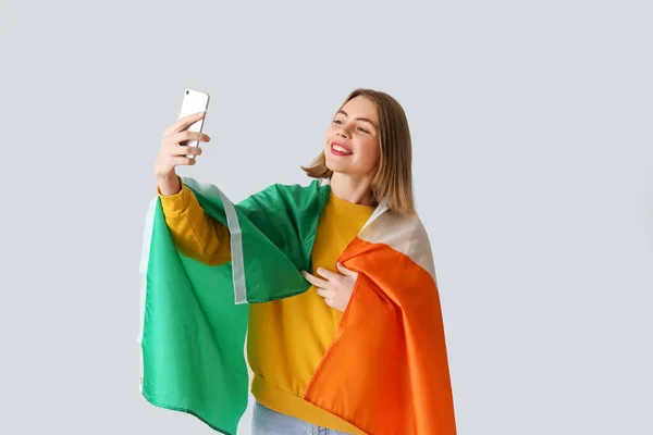 Young woman with flag of Ireland and mobile phone taking selfie on grey background