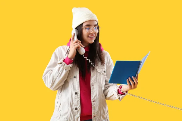 Pretty young woman with notebook talking by telephone on yellow background