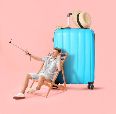 Genç erkek turist pembe arka planda selfie ve büyük bavul çekiyor.