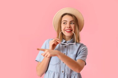 Şapkalı, güneş gözlüklü genç bir kadın pembe arka planda bir şeye bakıyor.