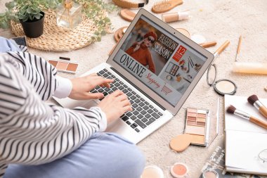 Dizüstü bilgisayarı, makyaj malzemeleri ve aksesuarları olan bir kadın bej halıda alışveriş yapıyor.