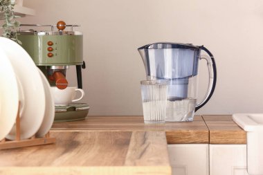 Bir bardak saf su ve ahşap mutfak tezgahında filtre sürahi.