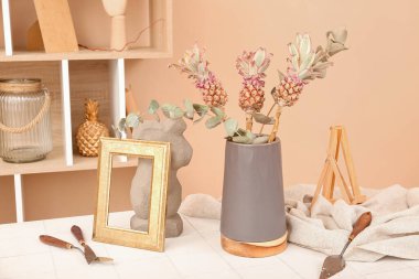 Süslü ananas vazoları, okaliptüs dalları, çerçeve ve spatulalar odadaki masada.