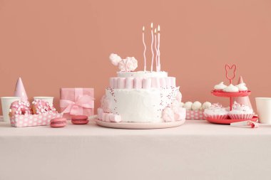 Koyu bej duvarın yanındaki beyaz masada mumlar ve şekerler olan doğum günü pastası.