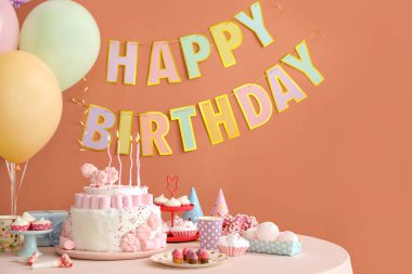 Koyu bej duvarın yanındaki beyaz masada mumlar ve şekerler olan doğum günü pastası.