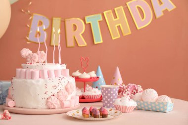 Koyu bej duvarın yanındaki beyaz masada mumlar ve şekerler olan doğum günü pastası.